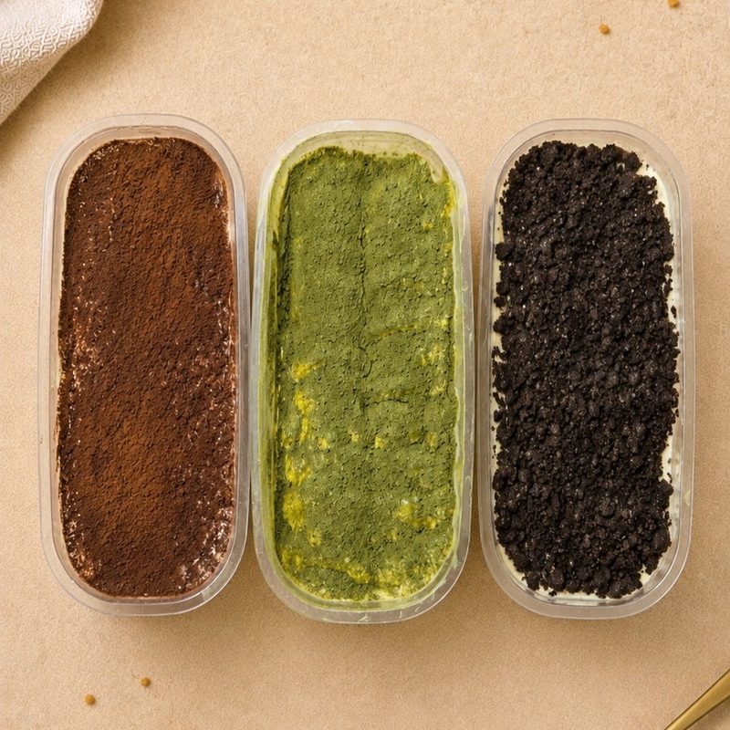 Three tubs of KRYM Delights tiramisu side by side &mdash; coffee dusted with cocoa, matcha, and Oreo crumb